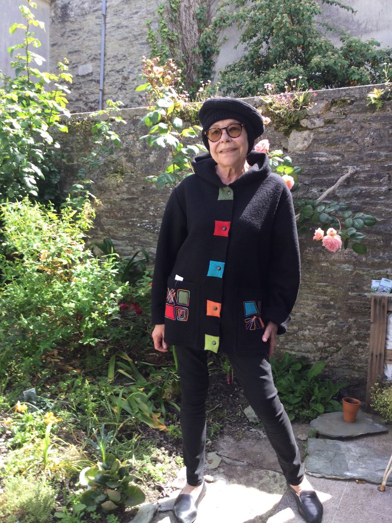 Une femme souriante avec un manteau noir et un beret noir, le tout agréments de patch de différentes couleurs vives..